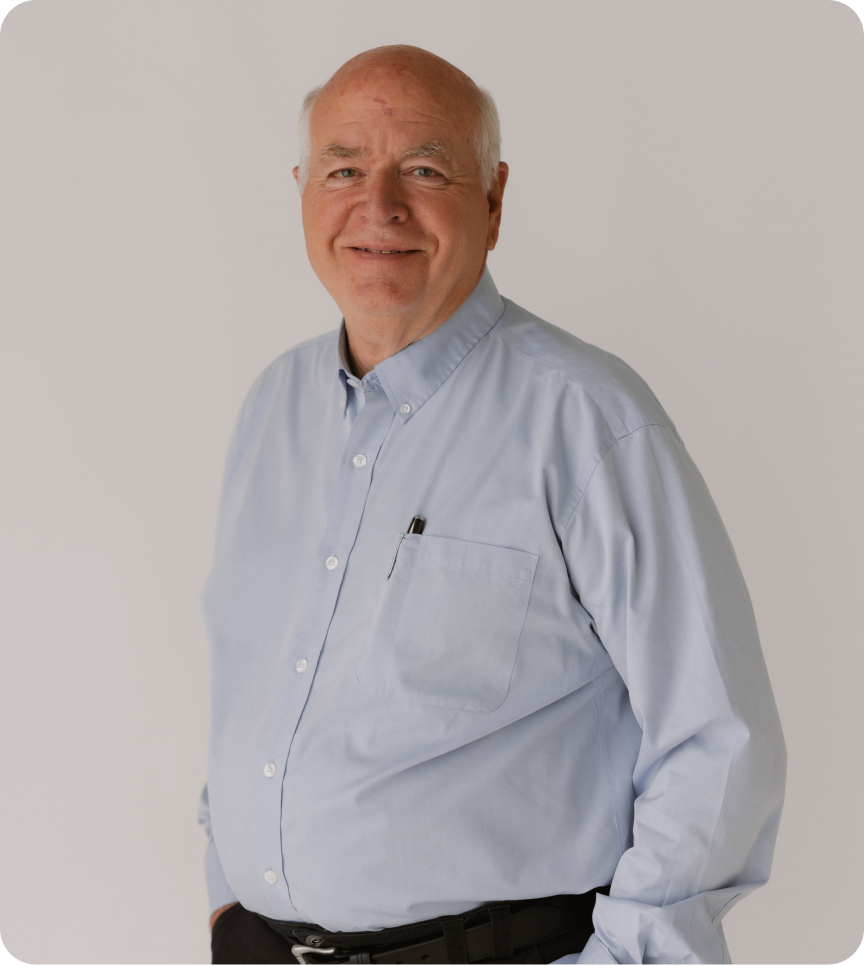 Older bald man in a light blue collared shirt and black pants standing in front of a white backdrop